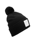A black knitted beanie hat features a bobble on top and a plain white rectangular patch on the front, shown against a white background.