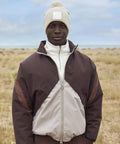 A person stands outdoors in a grass field wearing a brown and beige zip-up jacket, white polo neck, and the Beechfield Appliqué patch pom beanie, a beige knitted hat with a bobble. The sky is partly cloudy.