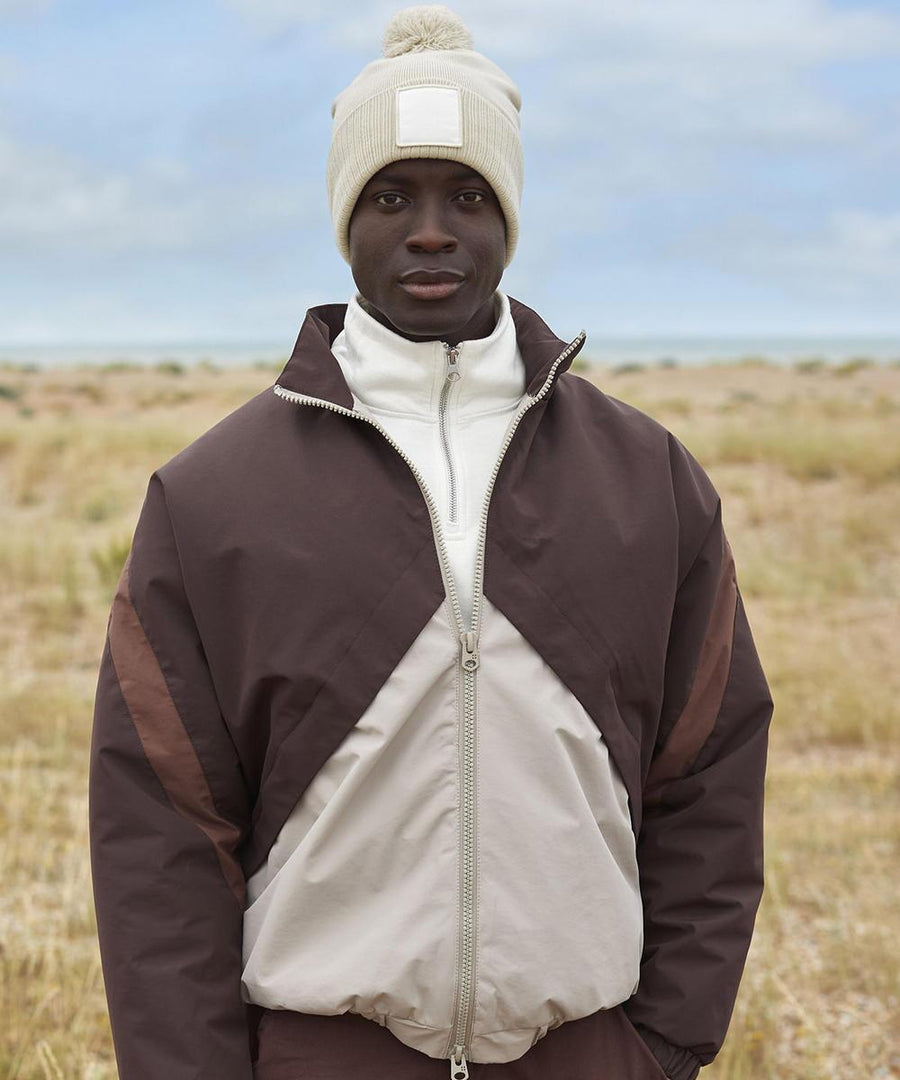 A person stands outdoors in a grass field wearing a brown and beige zip-up jacket, white polo neck, and the Beechfield Appliqué patch pom beanie, a beige knitted hat with a bobble. The sky is partly cloudy.