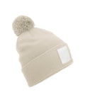 A beige knitted beanie hat with a pom-pom and a blank white rectangular patch on the turn-up, photographed against a white background.