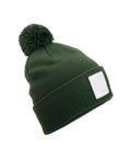 A dark green beanie knit hat features a pom-pom on top and a blank white rectangular patch on the folded brim, displayed against a white background.
