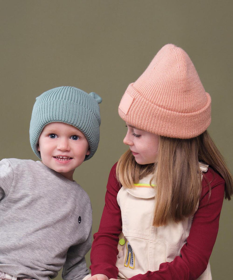 Two children wear Beechfield Organic baby bear beanies—one in light blue, the other in light pink—standing before an olive green backdrop. The child in blue smiles at the camera whilst the other looks at them.