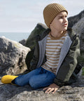 A boy wears the Beechfield Organic Cotton Kids Beanie, a brown knitted hat, as he sits on rocks by the water in a striped shirt and green jacket. He looks to the side with one yellow wellington boot resting nearby.