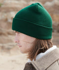 A person with shoulder-length hair wearing a green Beechfield B375R and a brown coat over a white turtleneck sweater is seen in profile. The background is outdoors with a blurred, natural setting.