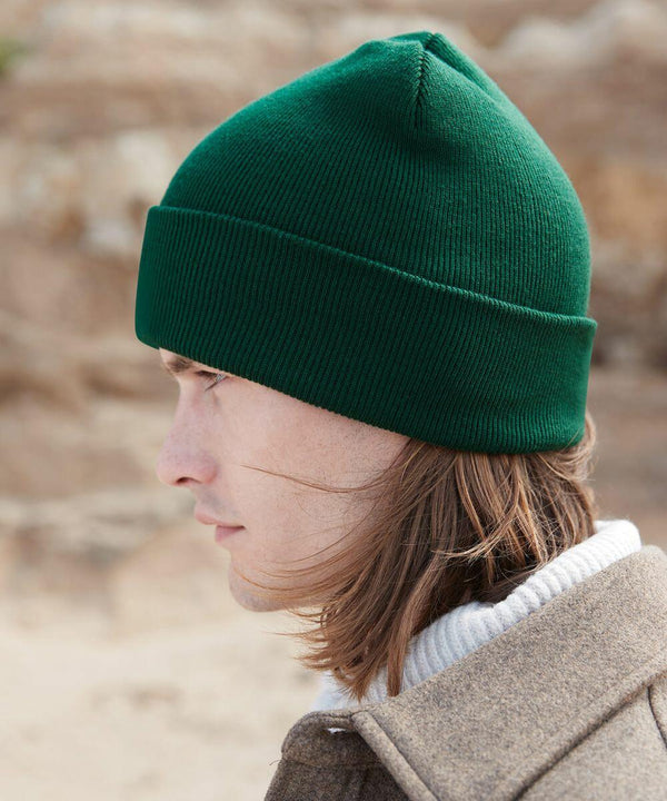A person with shoulder-length hair wearing a green Beechfield B375R and a brown coat over a white turtleneck sweater is seen in profile. The background is outdoors with a blurred, natural setting.