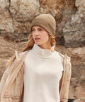 A woman with long blonde hair wearing a beige Beechfield B376R knit hat, off-white sweater, and tan jacket looks to the left against a rocky outdoor background.