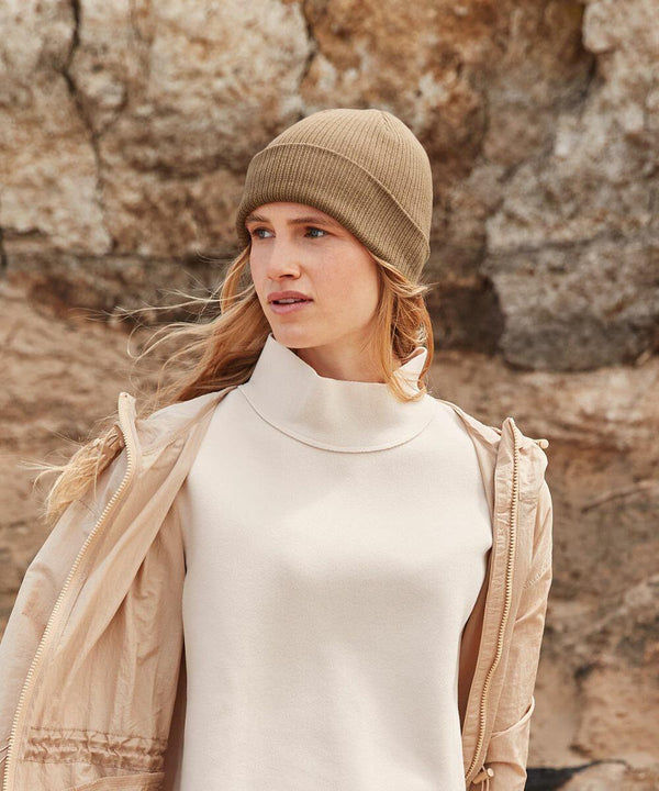 A woman with long blonde hair wearing a beige Beechfield B376R knit hat, off-white sweater, and tan jacket looks to the left against a rocky outdoor background.