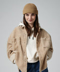 A person with long brown hair is smiling while wearing a tan knit beanie, a Beechfield B383R sherpa jacket, a beige hoodie, and blue jeans. They stand against a plain gray background with hands in their jacket pockets.