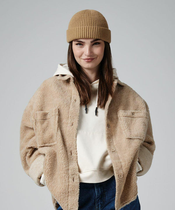 A person with long brown hair is smiling while wearing a tan knit beanie, a Beechfield B383R sherpa jacket, a beige hoodie, and blue jeans. They stand against a plain gray background with hands in their jacket pockets.