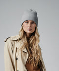 A person with long blonde hair wears a light gray knit Beechfield B384R beanie and a beige trench coat over a brown top. The background is plain and neutral.