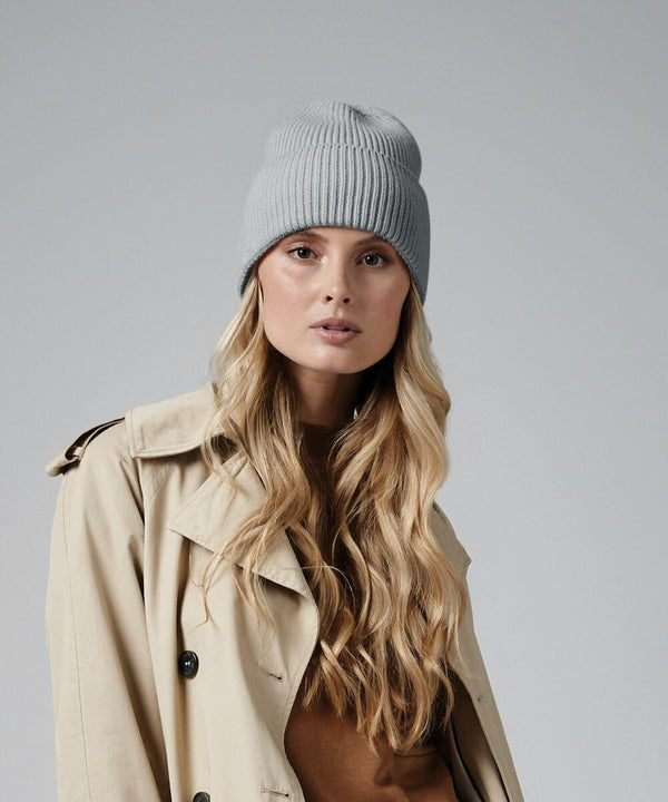 A person with long blonde hair wears a light gray knit Beechfield B384R beanie and a beige trench coat over a brown top. The background is plain and neutral.