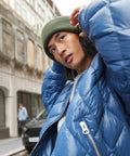 A person stands on a city street, wearing a blue puffer jacket and a Beechfield B385R. They are looking slightly upward with one hand adjusting the Beechfield B385R. The background shows buildings and a passing car.
