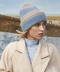 A person wearing a beige coat, light blue polo neck, and the Beechfield Colour Block Beanie stands outdoors near a weathered painted surface and grassy landscape.