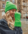A person with long, wavy blonde hair smiles while wearing a bright green Beechfield B397R knit hat and matching green knit sweater sleeve, covering part of their face. They are outdoors in a city with historic-looking buildings in the background.