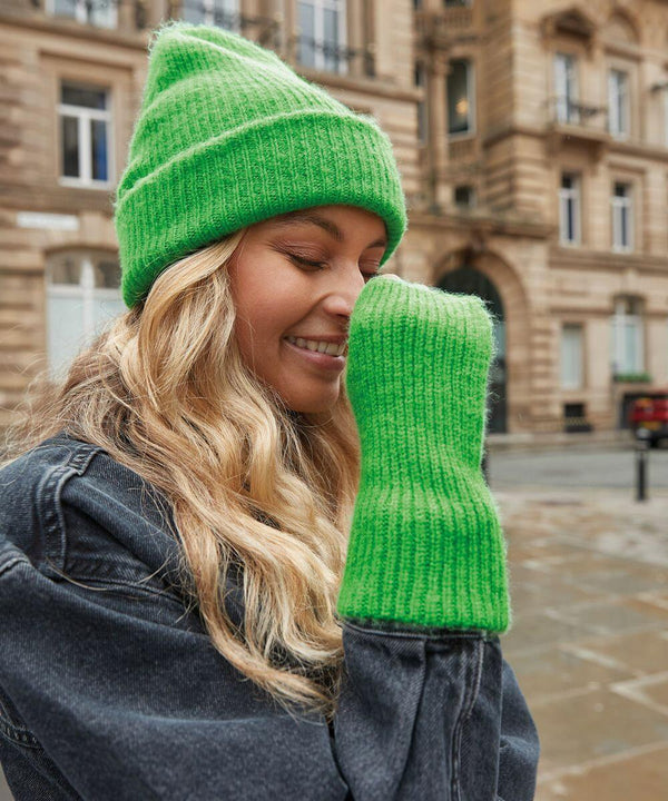 A person with long, wavy blonde hair smiles while wearing a bright green Beechfield B397R knit hat and matching green knit sweater sleeve, covering part of their face. They are outdoors in a city with historic-looking buildings in the background.