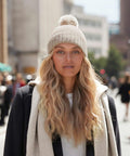 A person with long wavy blonde hair wearing a beige Beechfield B401R knit hat with a pom-pom, a white hoodie, and a dark coat stands outdoors. The background shows a city street with buildings and a few people walking.