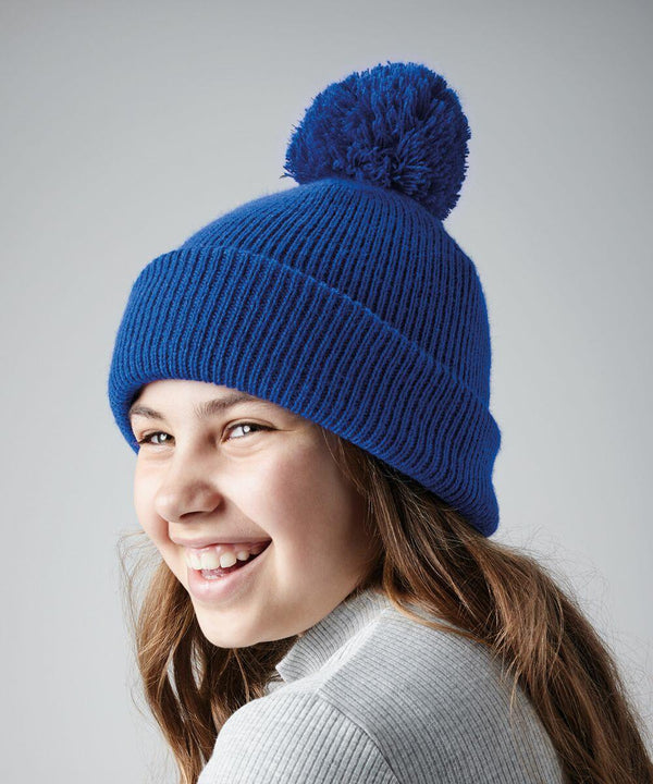 A smiling person with long hair, wearing a blue knit B406B beanie by Beechfield with a large pom-pom on top, and a light gray sweater. The background is a plain gray.