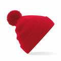 The Beechfield B426B beanie is a red knit hat featuring a large pom-pom on top. It is slightly tilted to the side, highlighting its ribbed texture and folded cuff.