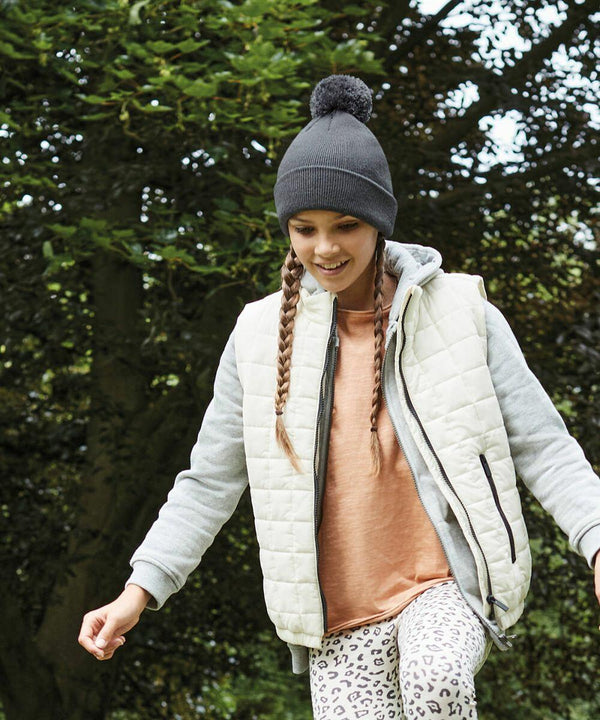 Smiling woman with long braids wearing a grey Beechfield B426B beanie, white quilted vest, grey hoodie, and peach shirt, walks outdoors. She is surrounded by green trees and appears to be enjoying a stroll in nature. Her leggings feature a leopard print pattern.