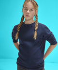 A young person with blond plaited hair wears the B&C Collection B&C ID.332 crew /kids dark blue long-sleeve shirt, standing against a bright blue background, hands behind their back, looking at the camera.