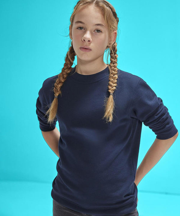 A young person with blond plaited hair wears the B&C Collection B&C ID.332 crew /kids dark blue long-sleeve shirt, standing against a bright blue background, hands behind their back, looking at the camera.