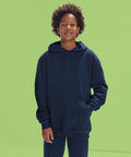 A boy with curly hair wears the B&C Collection B&C ID.333 hoodie/kids in dark blue with matching trousers, standing against a light green background. He has one hand in his front pocket and smiles slightly at the camera.