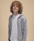 A young person with curly blond hair wears a B&C Collection B&C ID.334 zip hoodie /kids in light grey over a white T-shirt, standing before a plain beige background.