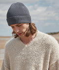 A person with shoulder-length hair wearing a gray knit Beechfield B438N and a cozy, textured, beige sweater stands outdoors on a beach. The sky is partly cloudy, and the person is looking downward with a serene expression.