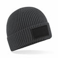 The Beechfield B442R is a dark gray beanie made with ribbed knit material and features a black rectangular patch on the front.