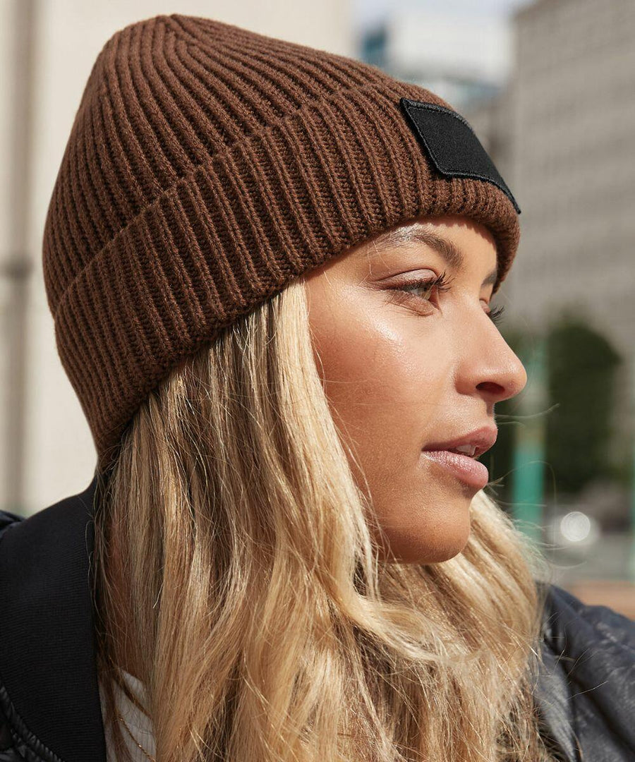 Close-up of a woman with long blonde hair wearing a Beechfield B442R ribbed brown beanie and black jacket, looking to the side. Urban buildings blurred in the background.