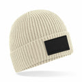 The Beechfield B442R is a beige ribbed knit beanie featuring a folded brim and a black rectangular patch on the side. The hat is shown against a white background.