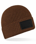 The B442R beanie from Beechfield features a brown ribbed knit design, a folded cuff, and a black rectangular patch on the front, all showcased against a white background.