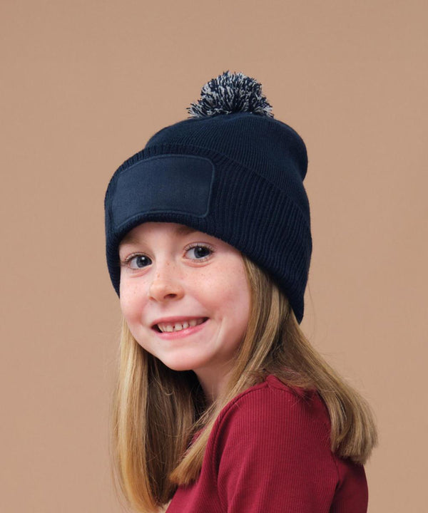 A young girl with light brown hair smiles whilst wearing the Beechfield Kids Snowstar Patch Beanie in navy blue, paired with a maroon long-sleeved shirt, against a plain beige background.