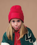 A young child with long blonde hair wears the Beechfield Kids’ Original Patch Beanie and looks directly at the camera, sticking out their tongue against a plain tan background.