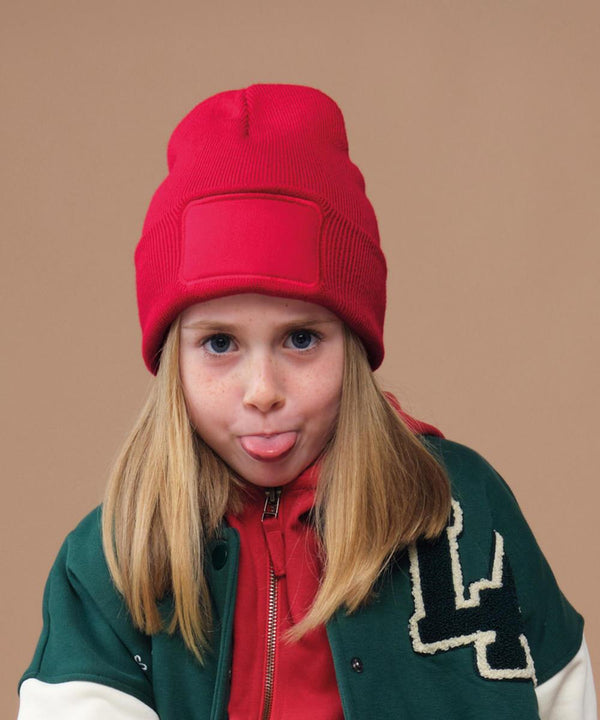 A young child with long blonde hair wears the Beechfield Kids’ Original Patch Beanie and looks directly at the camera, sticking out their tongue against a plain tan background.