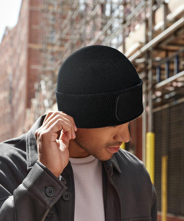 A person wearing a black Beechfield B445R beanie with a built-in headlamp is pictured adjusting the hat outdoors. They are dressed in a dark jacket and light shirt, standing in an urban area with buildings and scaffolding in the background on a sunny day.