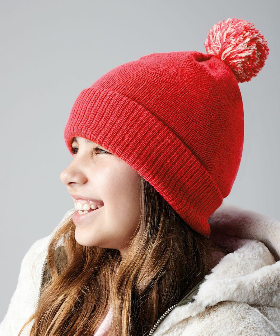 A young girl smiles while looking to the left. She is wearing a bright red Beechfield B450B knit beanie with a pom-pom on top and a white, fluffy jacket. The background is a plain, light gray color.