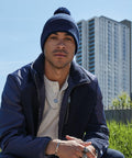 A person wearing a Beechfield B450N, a navy jacket with a shearling lining, and layered necklaces sits outdoors. A tall apartment building is visible in the background, along with some greenery. The sky is clear and blue.