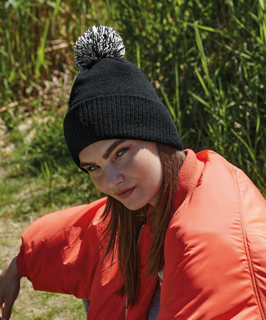 A woman wearing a black knit Beechfield B450R with a white and black pom-pom is sitting outdoors. She has on a bright orange jacket and has long brown hair. Green foliage can be seen in the background. The woman is looking slightly to the side with a subtle smile.
