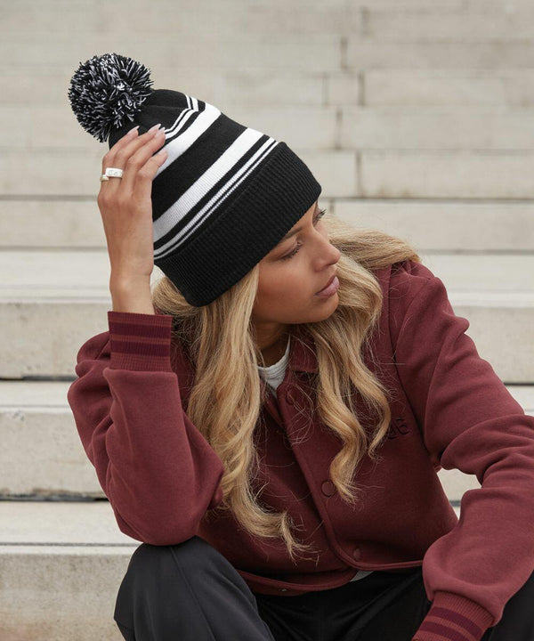 Person with long blonde hair sitting on steps, wearing a maroon jacket and a Beechfield B473R beanie with a pom-pom. They are resting their head on one hand, appearing thoughtful.