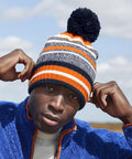 A person wearing a blue fleece jacket and a Beechfield Multi-sport fan beanie featuring orange, white, grey, and black stripes adjusts their hat outdoors on a partly cloudy day.