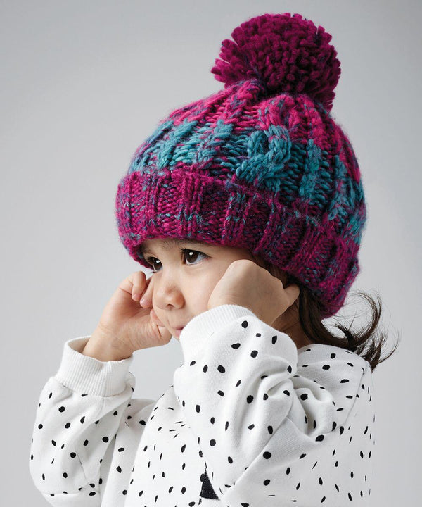 A young child with brown hair wears a polka-dotted shirt and a colorful knitted hat with a pom-pom. The hat, Beechfield's B486A, has a blend of pink, purple, and turquoise colors. The child is looking to the side with their hands near their face. The background is plain gray.