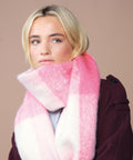 A person with blonde hair wears a large Beechfield fluffy check scarf in pink and white check and a dark maroon coat, looking slightly to the side against a plain beige background.