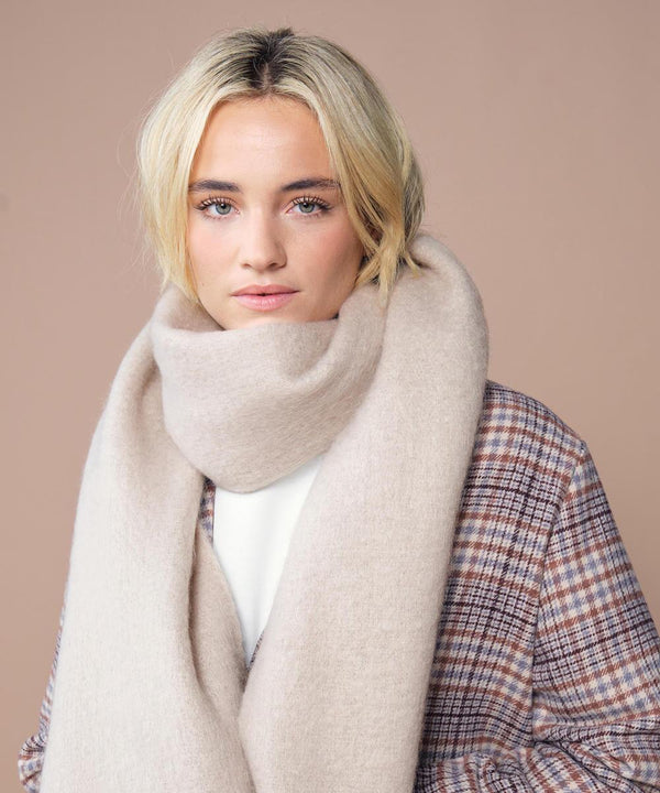 A woman with blonde hair models the Beechfield Fluffy scarf, styled in beige, paired with a checked coat and white top. She stands against a light brown backdrop, gazing neutrally at the camera.
