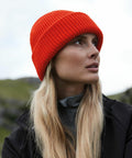 A person with long blonde hair wearing a Beechfield B508R looks off to the side. They are dressed in a dark jacket with a grey zip-up underneath. The background is slightly blurred, showing hints of a natural landscape with greenery.