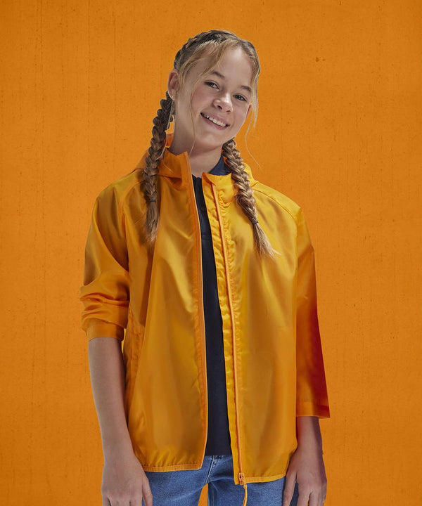 A young person with plaited hair wears the B&C Collection B&C #Reset windcheater /kids in bright orange over a dark shirt and blue jeans, standing against an orange background and smiling at the camera.
