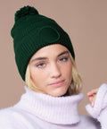 A person wearing the Beechfield Fan patch beanie in green and a light purple polo neck jumper looks at the camera against a plain beige background.