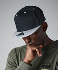 A man wearing a black and white Beechfield B610C snapback cap and an olive green shirt with globe graphic is resting his chin on his hand, looking downward pensively against a plain gray background.