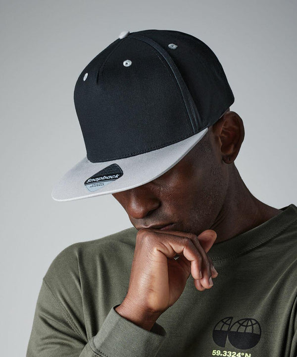 A man wearing a black and white Beechfield B610C snapback cap and an olive green shirt with globe graphic is resting his chin on his hand, looking downward pensively against a plain gray background.