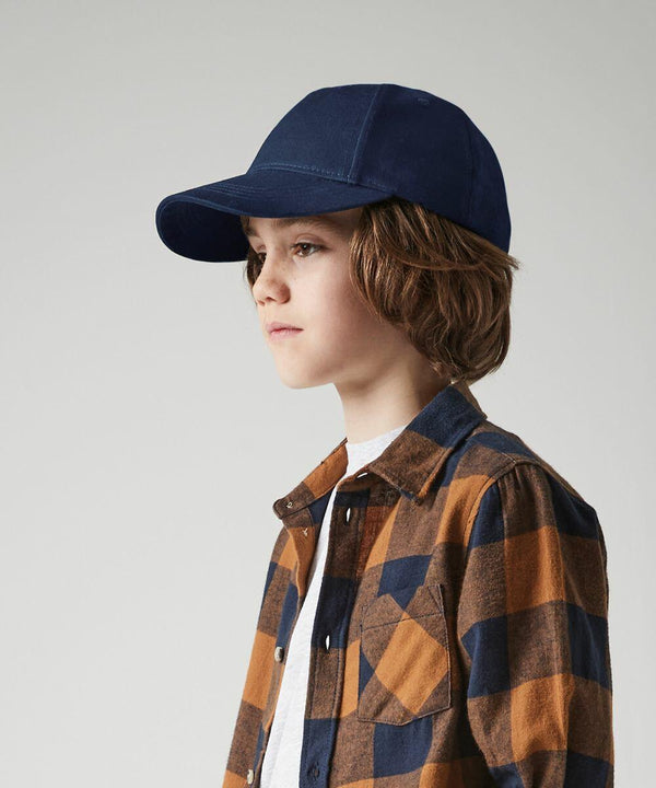 A young person with medium-length hair is wearing a blue Beechfield B63NB cap and a brown plaid shirt over a white t-shirt. They are facing to the right with a neutral expression against a plain background.